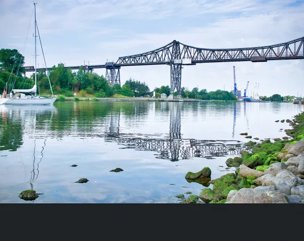 Eisenbahn-Hochbrücke über den Nord-Ostsee-Kanal in Rendsburg, daneben ein Segelboot auf dem Wasser.