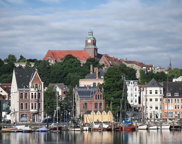 Hafen von Schleswig-Flensburg mit bunten Gebäuden und Booten vor einer grünen Hügelkulisse.