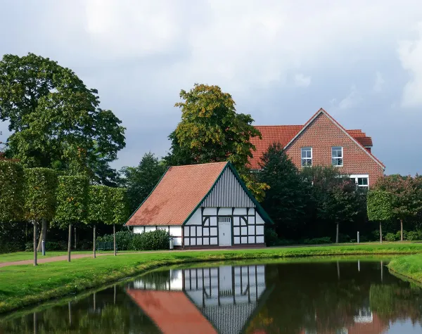 Fachwerkhaus mit roten Dachziegeln am Teich, umgeben von Bäumen und Wiesen in Diepholz.