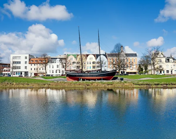 Segelschiff vor historischer Gebäudezeile in Cuxhaven, reflektierend im Wasser bei blauem Himmel.