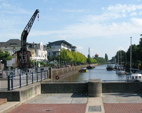Wasserweg in Oldenburg mit Promenade, Booten und altem Kran, flankiert von modernen und historischen Gebäuden.