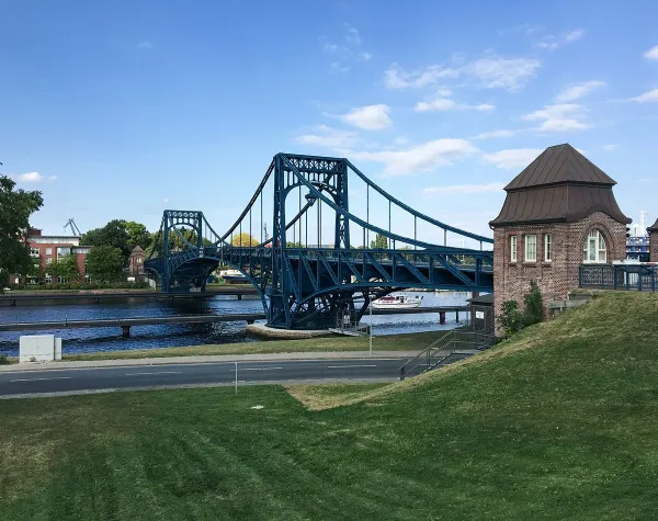Blaue Eisenbahnbrücke über einen Fluss in Wilhelmshaven, mit historischem Gebäude im Vordergrund und blauem Himmel.