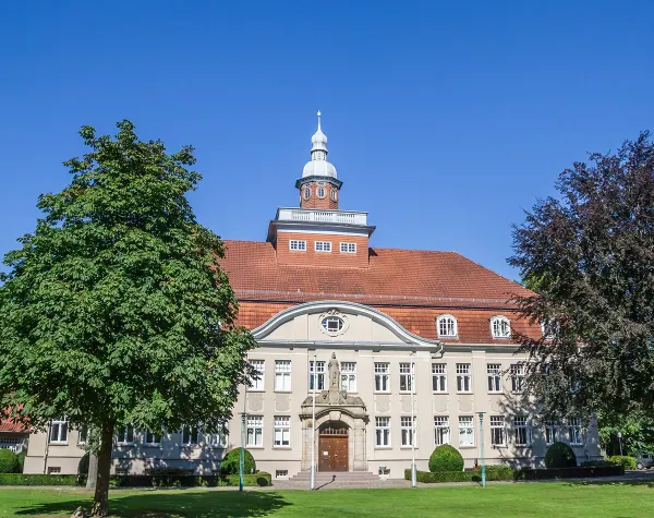 Historisches Gebäude in Cloppenburg mit rotem Dach, Uhrturm und symmetrischen Fenstern, umgeben von grünen Bäumen.