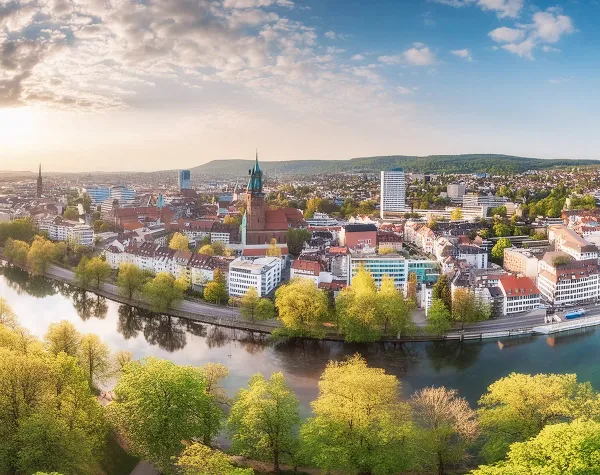 Panorama der Stadt Bielefeld mit Altstadt, Kirchen und Fluss, umgeben von grüner Landschaft unter bewölktem Himmel.