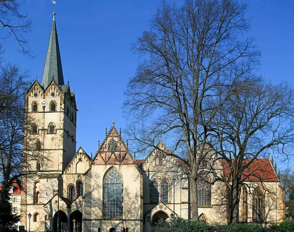 Kirche in Herford mit gotischer Architektur und hohen Bäumen im Vordergrund unter blauem Himmel.