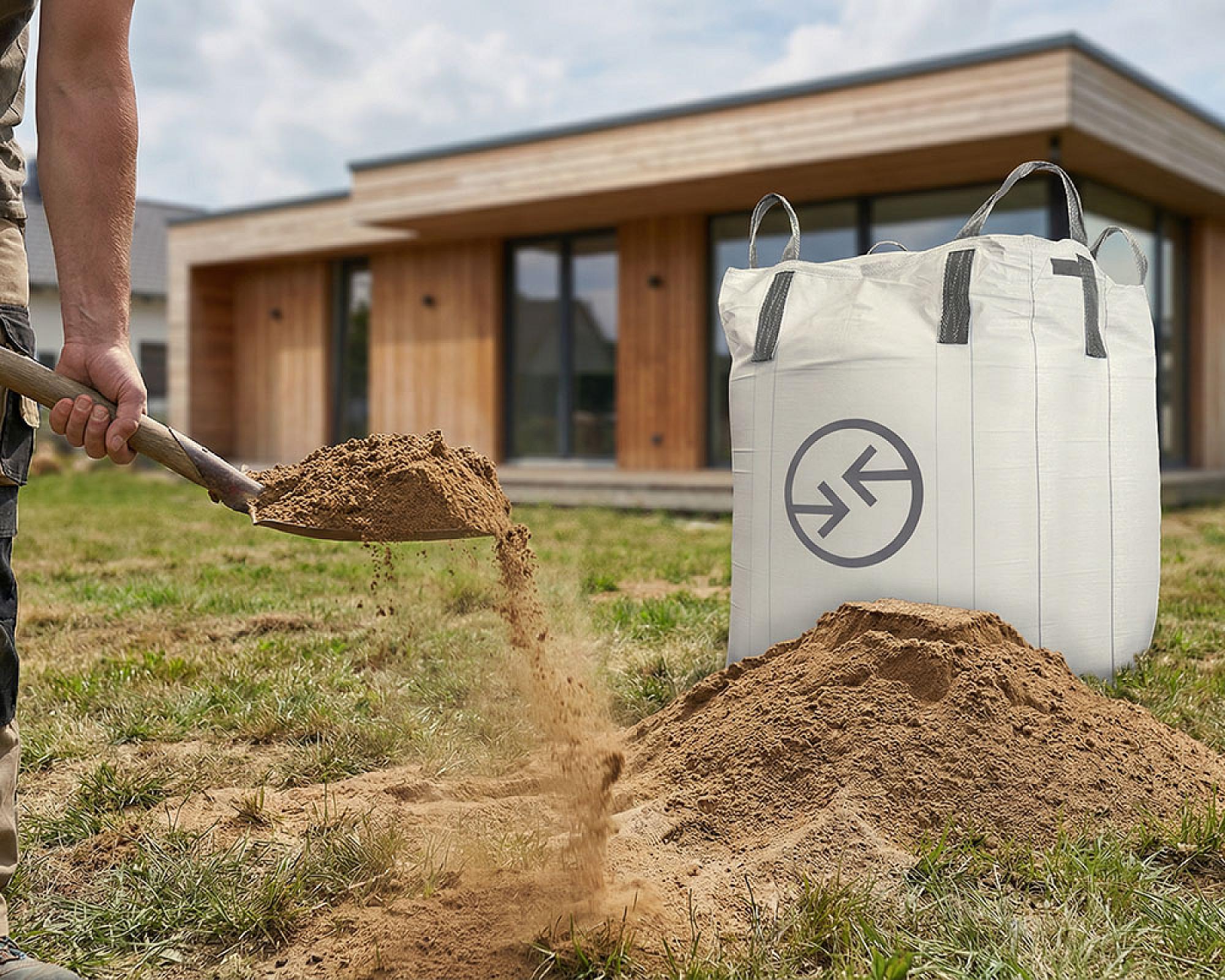 Person schaufelt Rasensand aus einem großen Big Bag vor einem modernen Holzhaus.