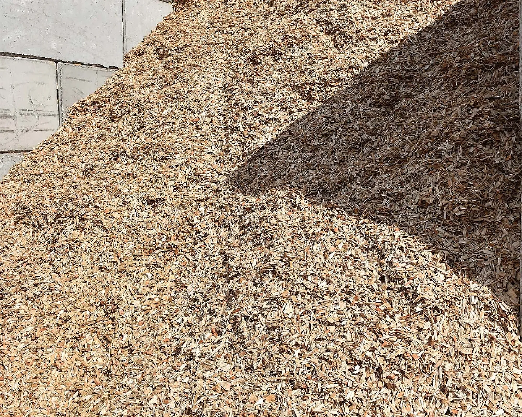 Lose Holzhackschnitzel in einem großen Haufen auf einer Betonfläche. Schatten fällt teilweise über den Haufen.