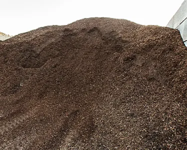 Ein großer Haufen feiner, brauner Rindenmulch auf Erde vor einer hellen Wand.