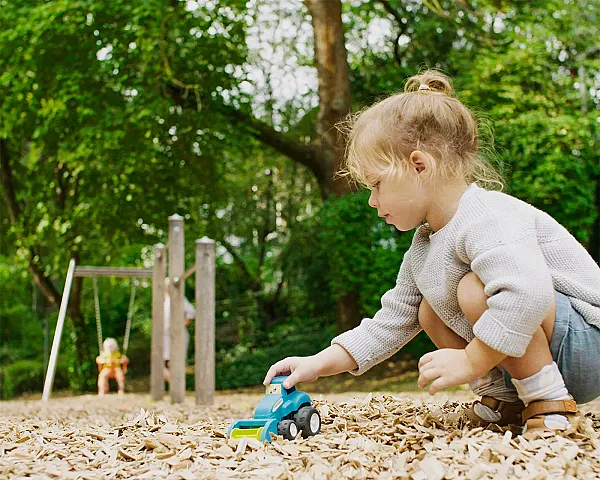 Kind spielt mit Spielzeugauto auf Fallschutz-Hackschnitzel-Untergrund, im Hintergrund eine Schaukel im Park.