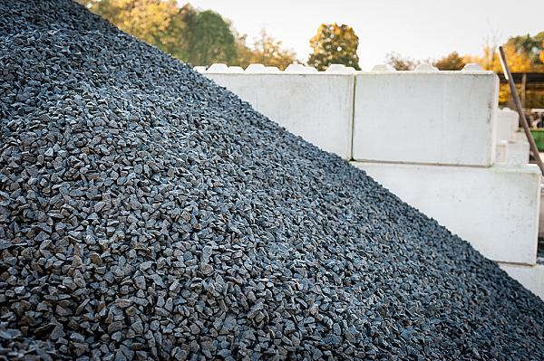 Dunkelgrauer Basaltsplitt aufgetürmt vor hellen Betonblöcken im Außenbereich.