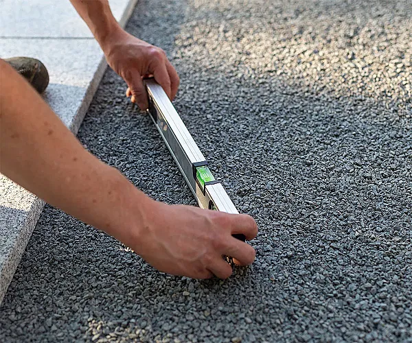 Person nivelliert grauen Pflastersplitt mit Wasserwaage auf Baustelle.