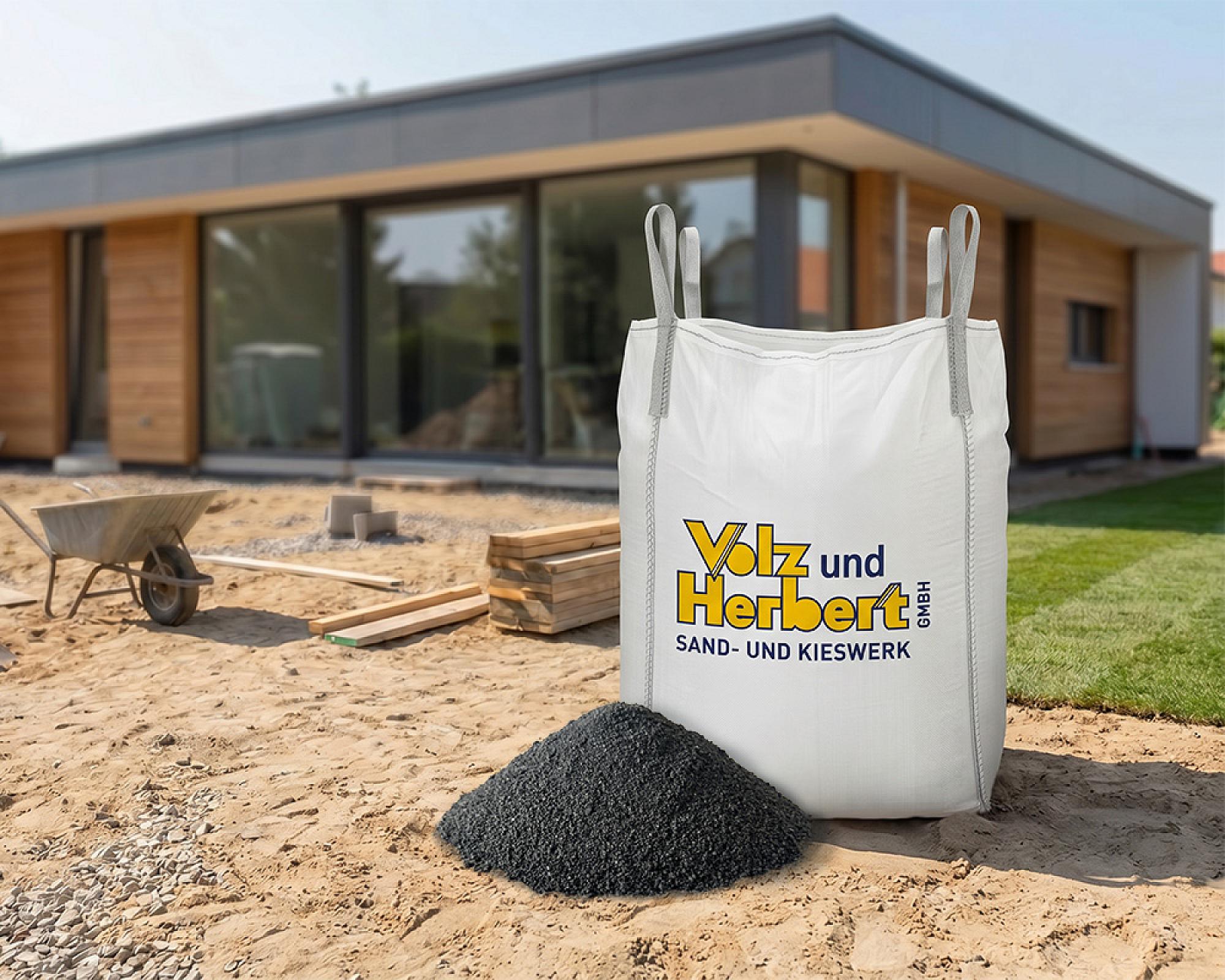 Großer Big-Bag mit Basaltsand vor modernem Gebäude auf Baustelle, daneben Schubkarre und Holzstapel.