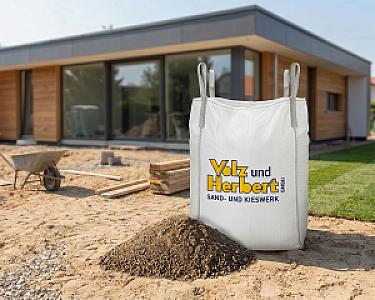Big Bag mit Estrichsand vor modernem Haus auf Baustelle, daneben Schubkarre und Holzstapel.