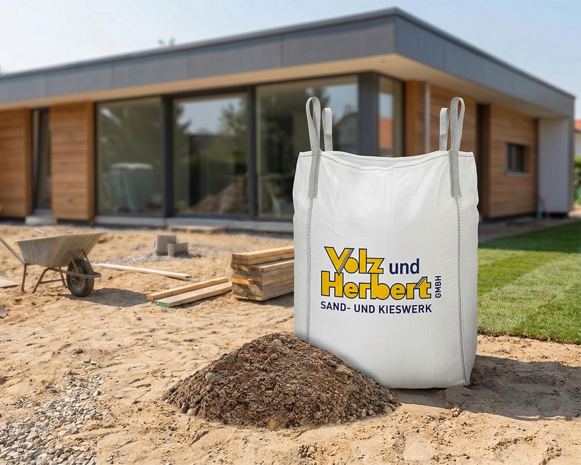 Bigbag Betonkies auf Baustelle vor modernem Gebäude, daneben Schubkarre und Holzstapel.