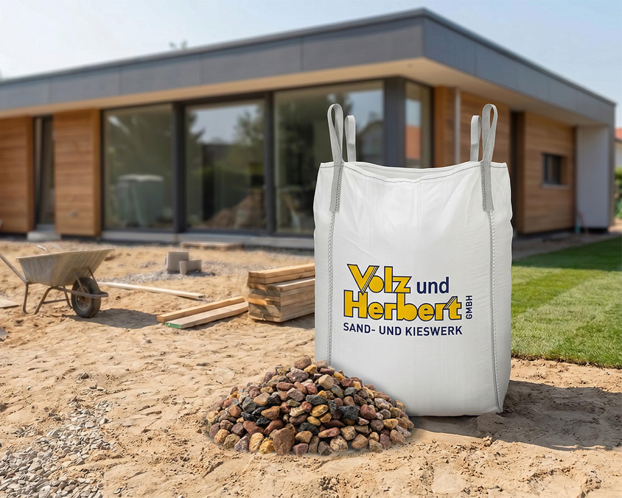 Großer Big Bag mit Drainagekies der Marke Volz und Herbert GmbH auf Baustelle vor modernem Holzhaus.