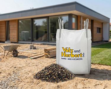 Großer Bigbag mit Volz und Herbert Logo, gefüllt mit Fallschutzkies, vor einem modernen Gebäude auf Baustelle.