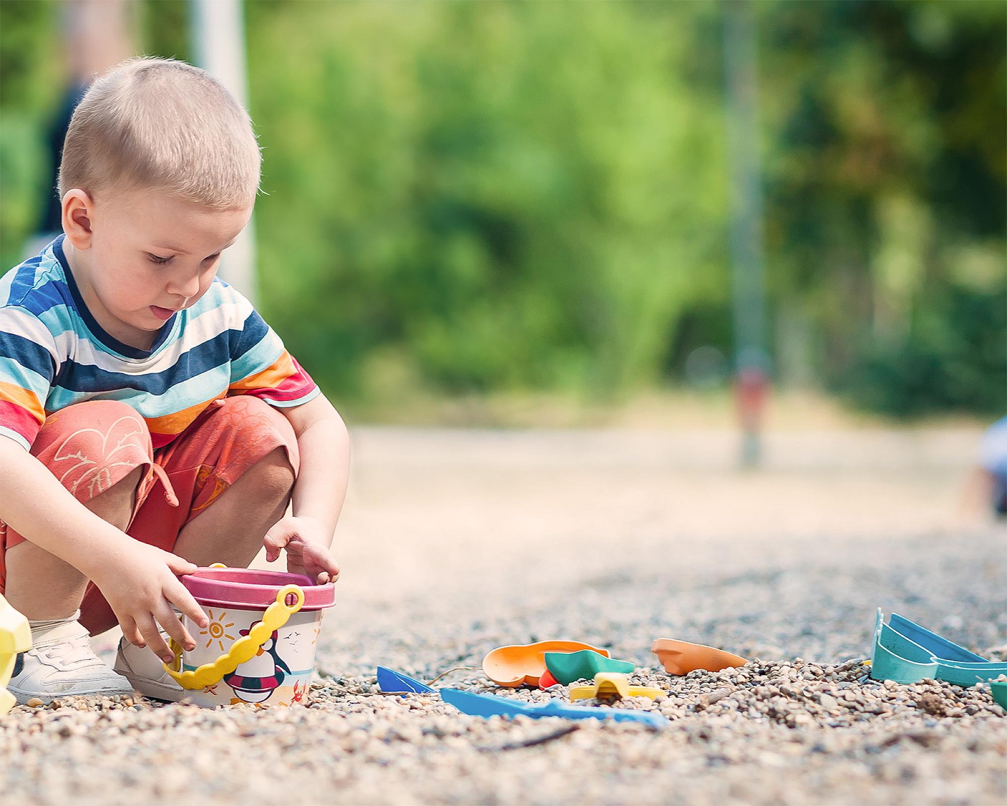 Kind spielt mit buntem Spielzeug im Fallschutzkies, sommerliche Umgebung, Fokus auf Sicherheit und Spielvergnügen.