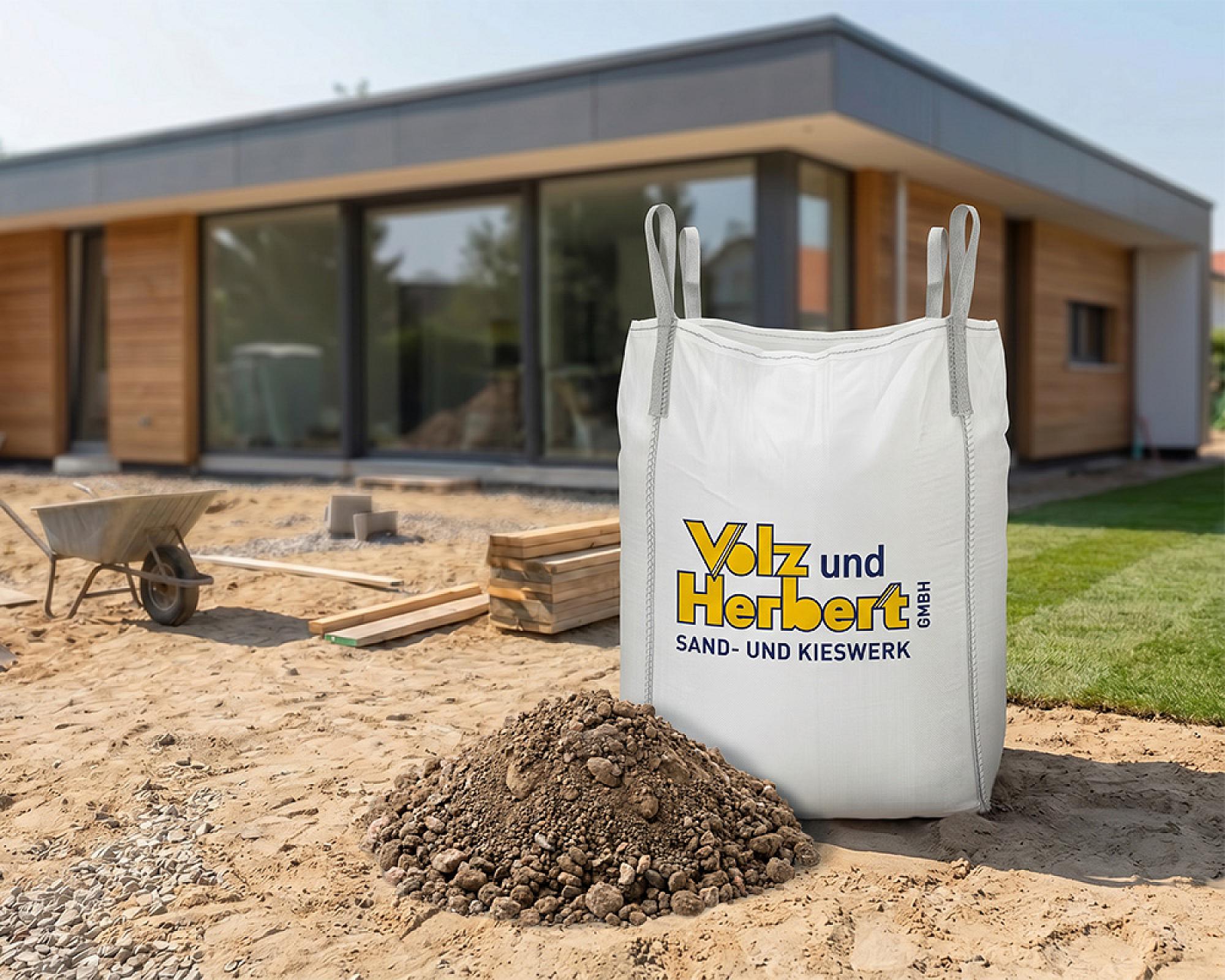 Großer Big Bag mit Logo von Volz und Herbert GmbH, gefüllt mit Füllkies, auf Baustelle mit modernem Haus im Hintergrund.