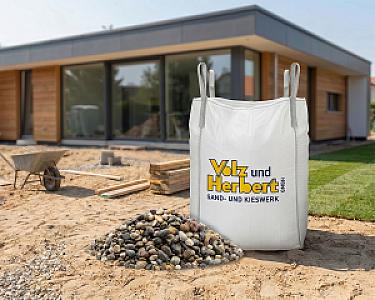 Großer Big Bag mit Rheinkies vor modernem Holzhaus, daneben Schubkarre auf Baustelle.