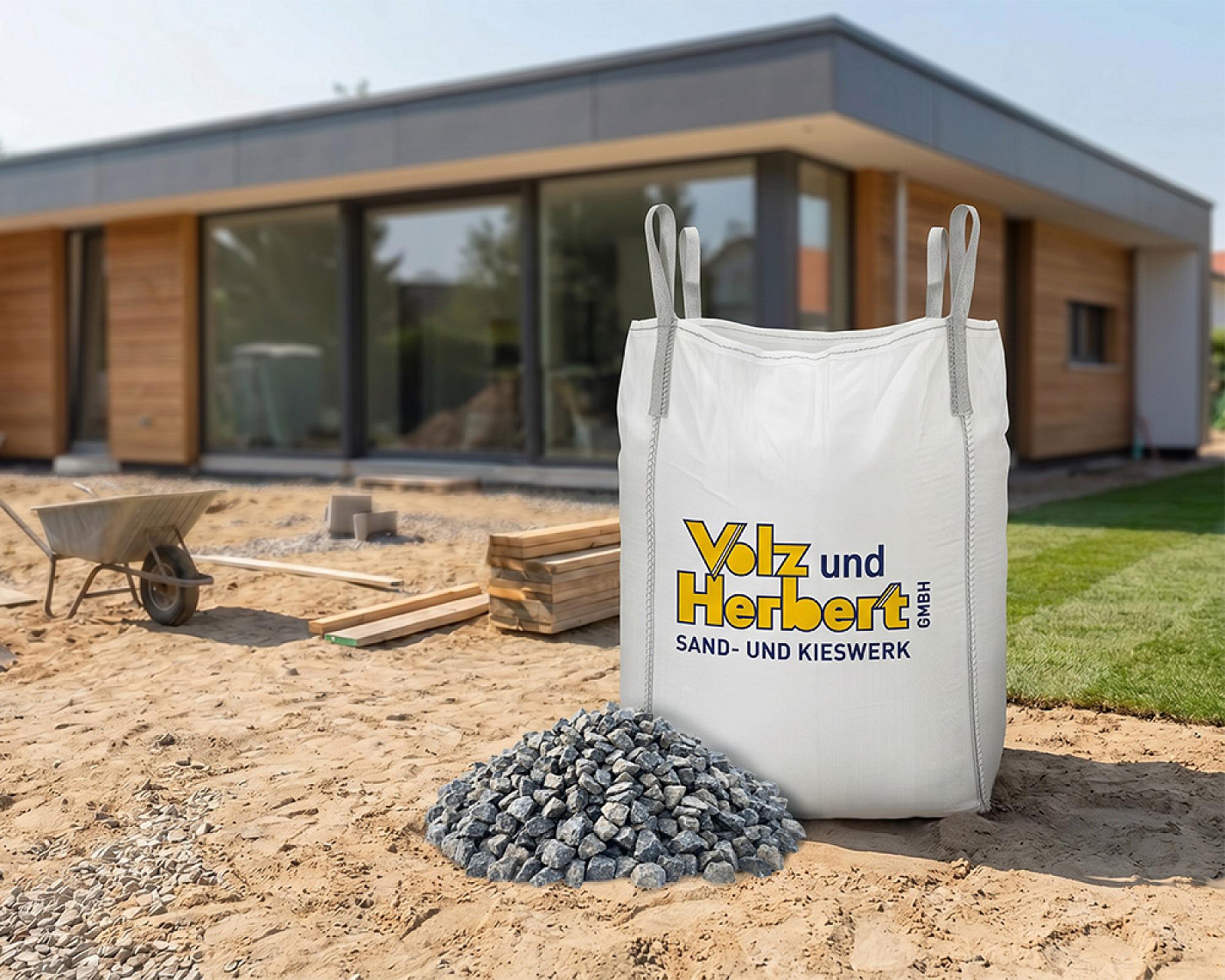 Großer weißer Big Bag mit Volz und Herbert-Logo, gefüllt mit Basaltsplitt, auf Baustelle vor moderner Holzkonstruktion.