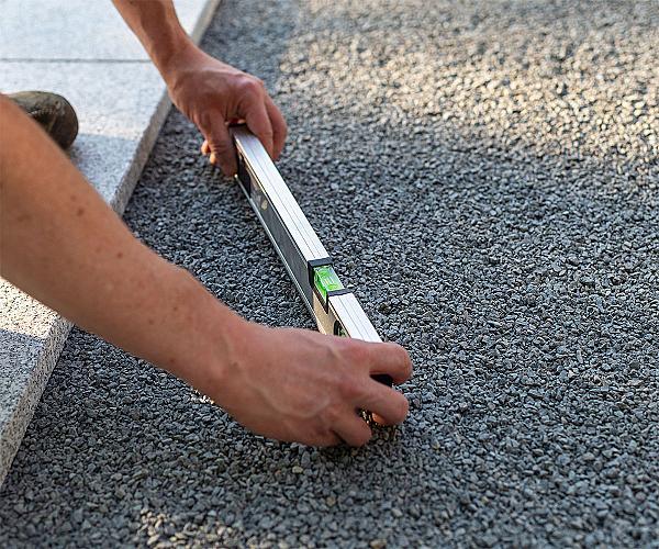 Hände richten mit einer Wasserwaage eine Schicht Pflastersplitt auf einer Baustelle aus.