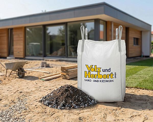 Großer Bigbag mit Aufschrift vor modernem Haus, gefüllt mit Vegetationsschicht; Sand und Kies im Vordergrund.
