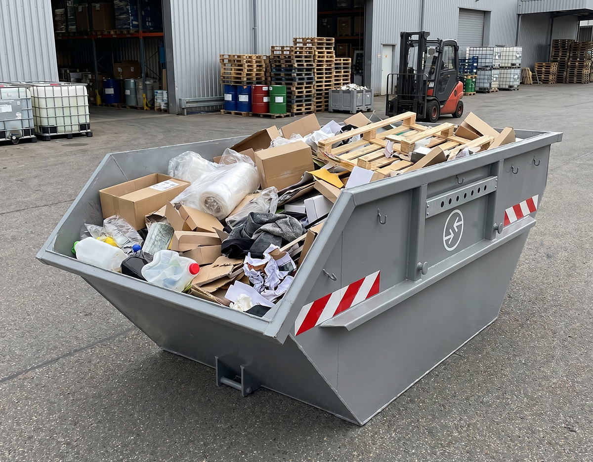 Gewerbeabfall-Container aus Metall, gefüllt mit Kartons und Verpackungsmaterial, auf Industriegelände.