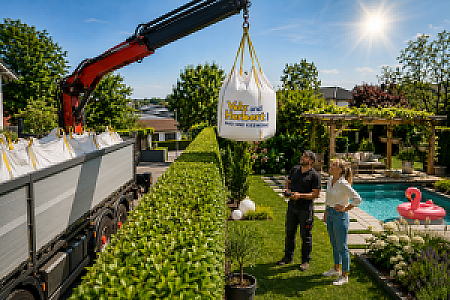 Kran hebt BigBag mit Felsenkies von Volz und Herbert GmbH in Garten, sichtbarer Pool und Paar im Gespräch.