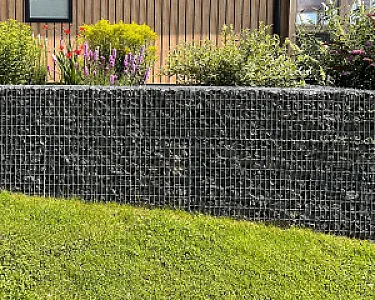 Gabionenzaun mit anthrazitfarbenen Basaltsteinen, flankiert von grünen Pflanzen und blühenden Blumen im Garten.