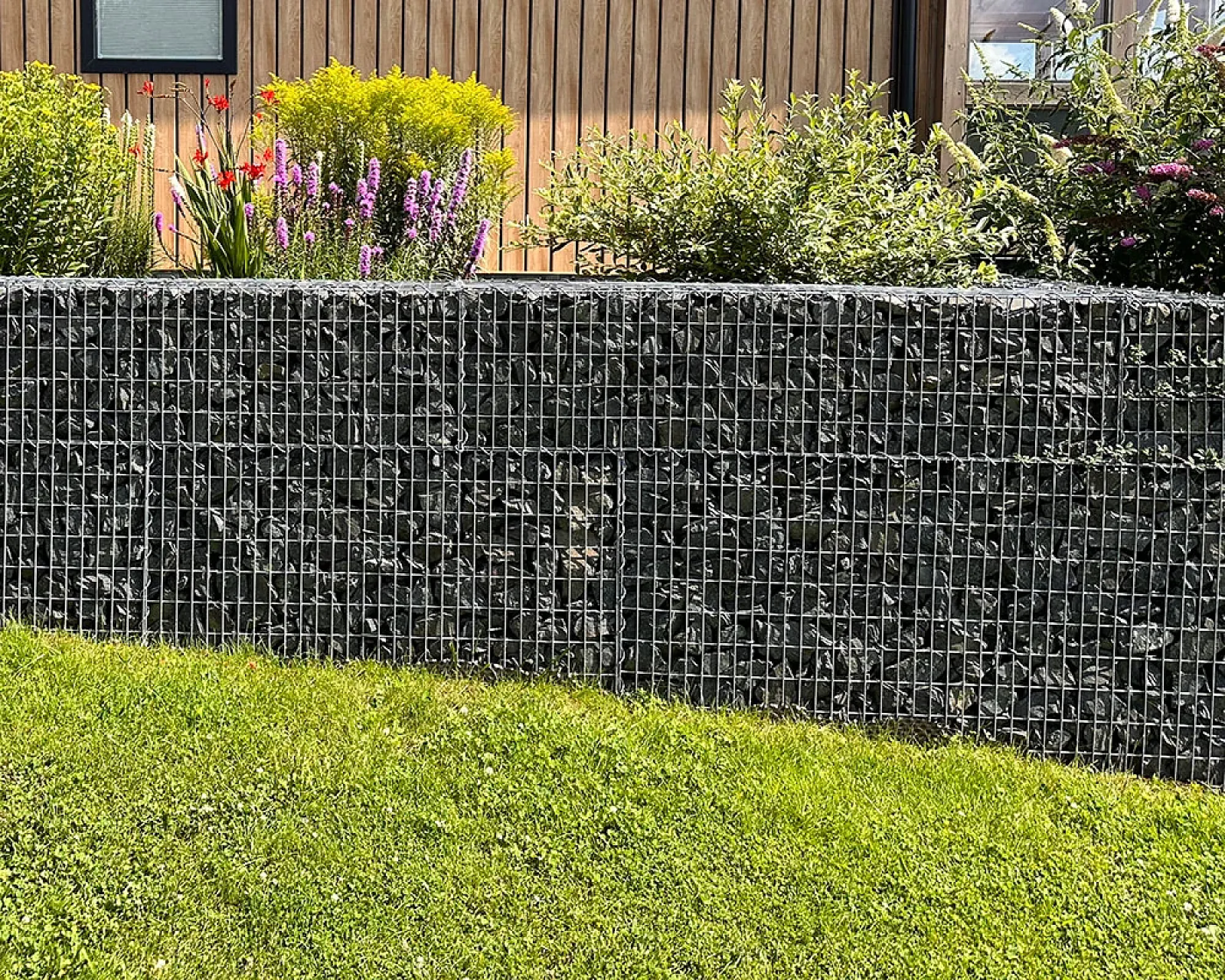 Gabionenwand gefüllt mit anthrazitfarbenen Basaltsteinen umgeben von grüner Gartenlandschaft.