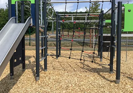 Spielplatz mit Klettergerüst und Fallschutz aus Hackschnitzeln; umgeben von grüner Natur.