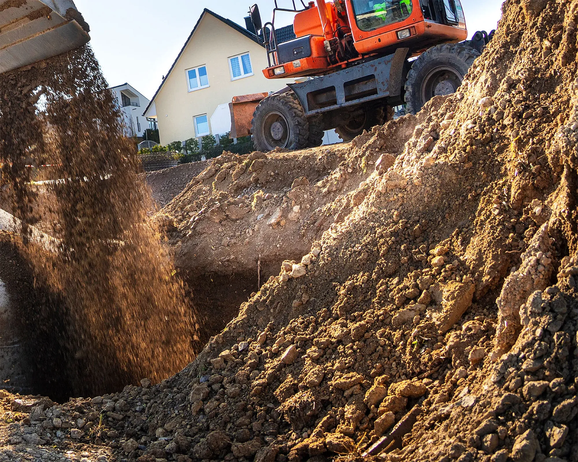 Bagger bei Erdarbeiten bewegt Füllboden; Bauschutt sichtbar, ländliche Umgebung mit Wohnhaus im Hintergrund.