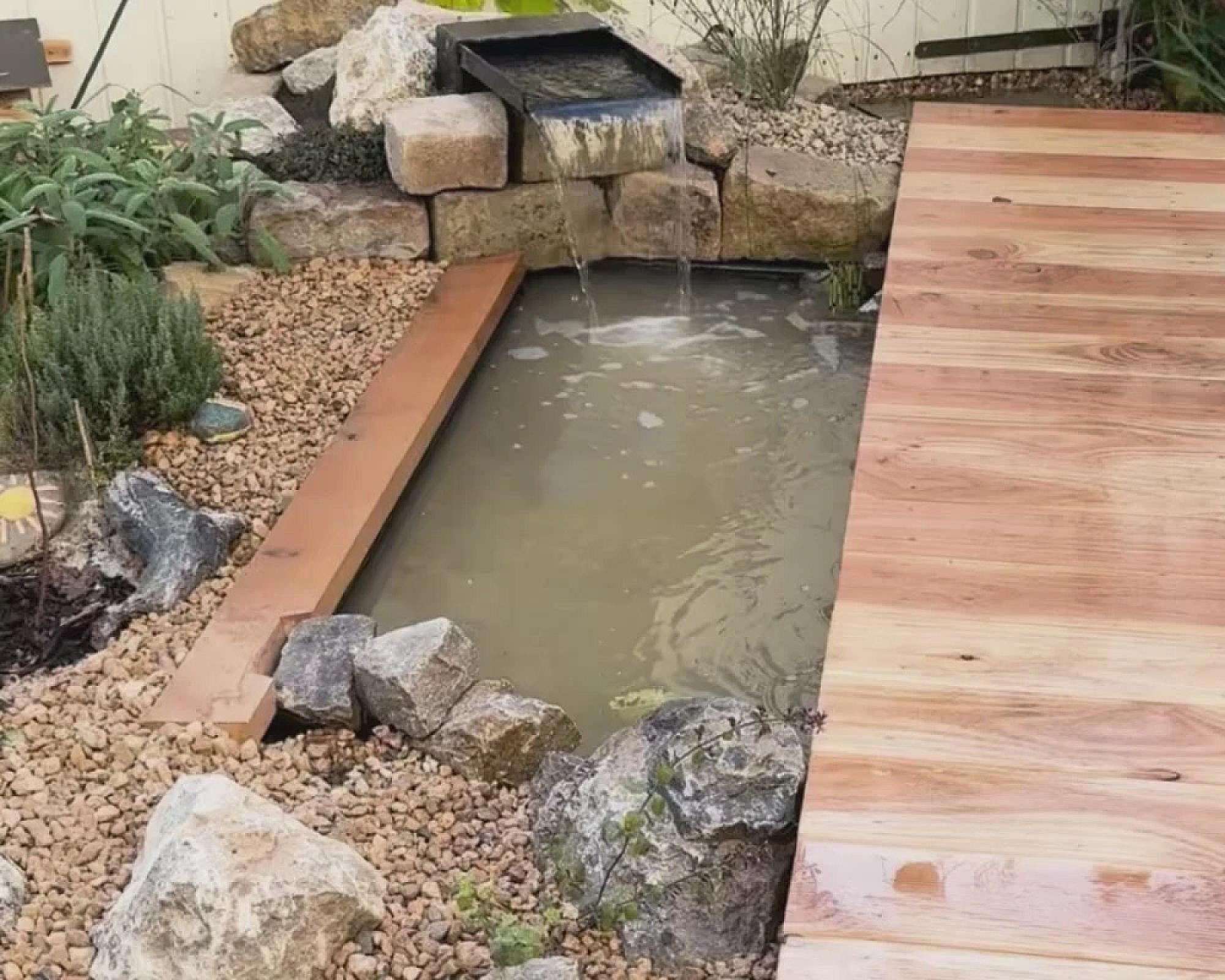 Steinbereich mit Drainagekies ums Gartenteich, hölzernes Deck, Bachlauf im Hintergrund.