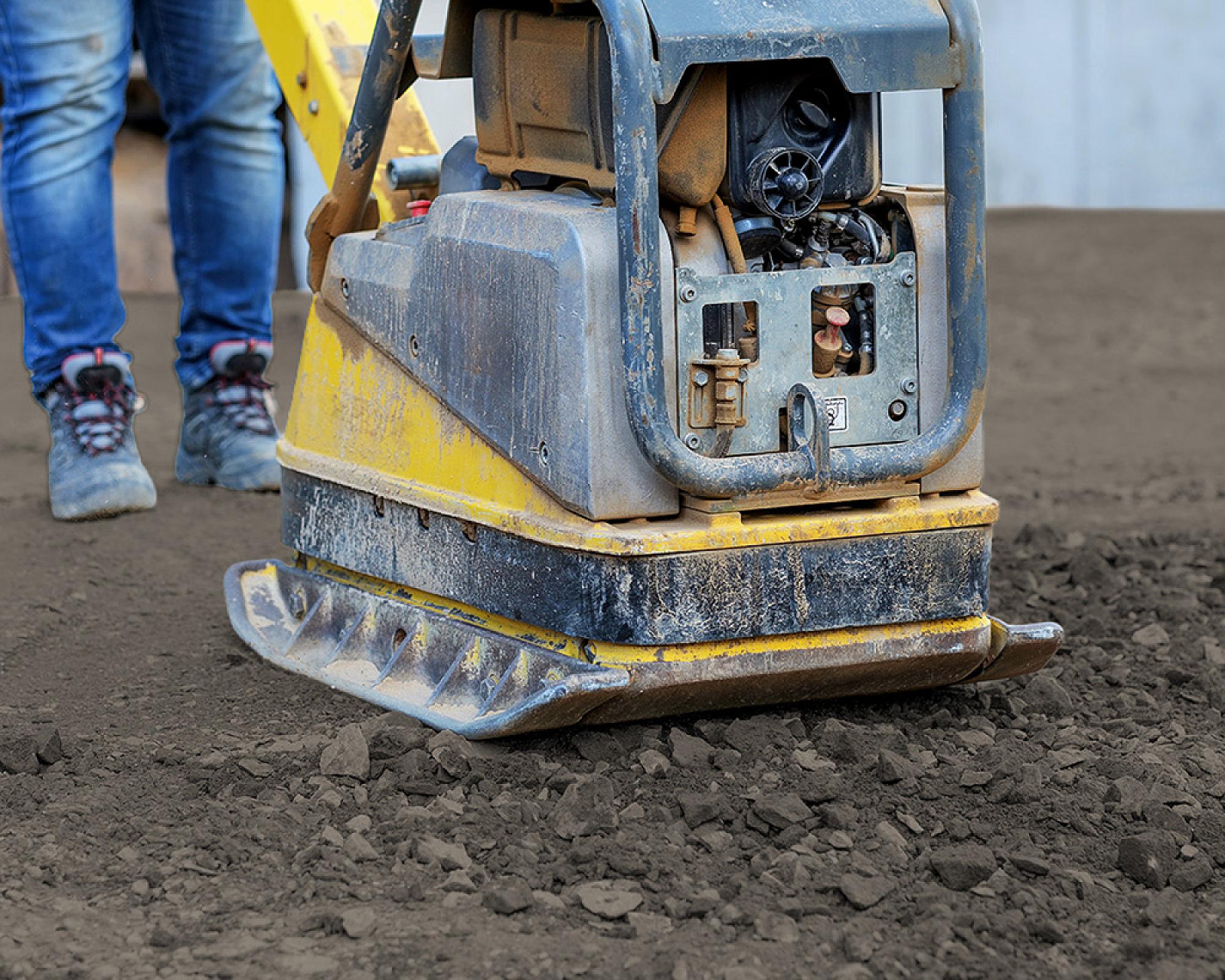Baumaschine verdichtet braunes Mineralgemisch, Arbeiter trägt Jeans und Sicherheitsschuhe im Hintergrund.