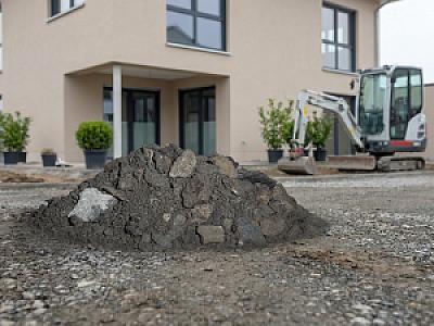 Mineralgemisch vor modernem Gebäude, Hintergrund: kleiner Bagger, Pflanzen in Töpfen.