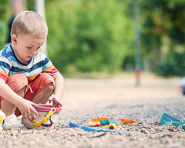 Kind spielt mit buntem Eimer auf einem Boden aus Fallschutzkies, umgeben von Sandspielzeug bei sonnigem Wetter.