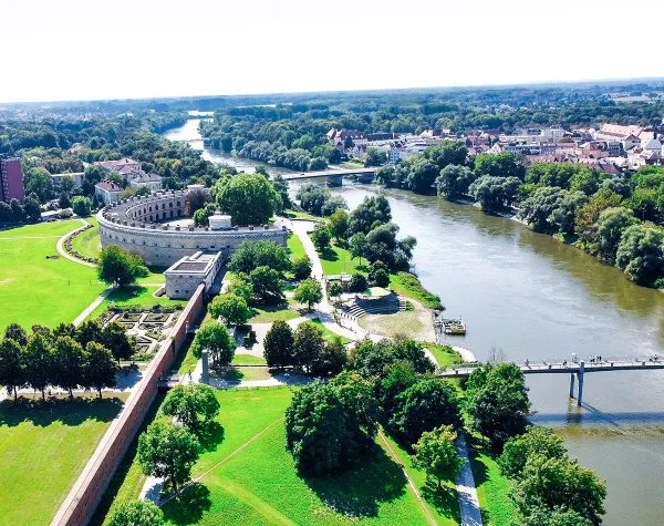 Blick auf Ingolstadt mit Fluss, Grünflächen und historischen Gebäuden bei sonnigem Wetter.