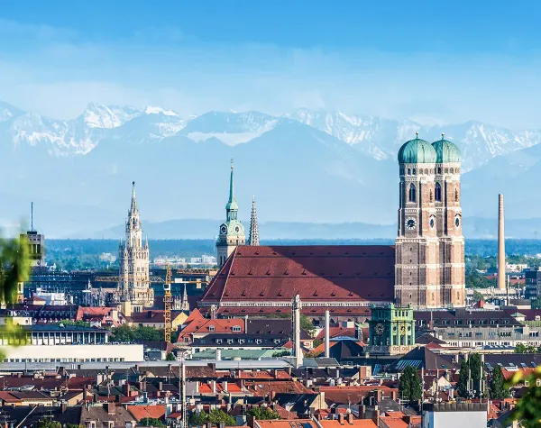 Münchens Skyline mit Frauenkirche und Alpen im Hintergrund unter blauem Himmel.