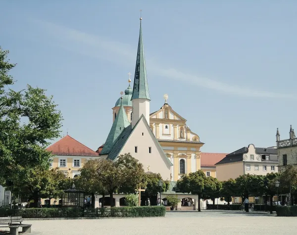 Pilgerkirche in Altötting, Deutschland, mit markantem Kirchturm und umliegenden Gebäuden auf einem sonnigen Platz.