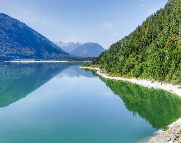 Klarer See in alpiner Landschaft bei Bad Tölz-Wolfratshausen, umgeben von bewaldeten Bergen und blauem Himmel.