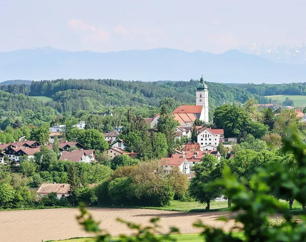 Landschaftsansicht von Ebersberg mit Kirche und umliegenden Häusern, im Hintergrund bewaldete Hügel und Berge.