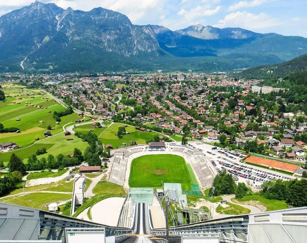 Blick von Skischanze auf Garmisch-Partenkirchen, umgeben von Bergen, mit Grünflächen und Wohnhäusern im Tal.