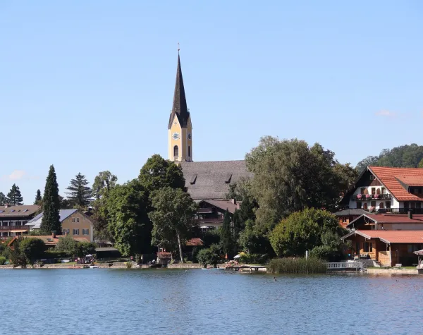 Kirche mit hohem Turm und Dorfhäusern am Ufer eines Sees in Miesbach bei klarem Himmel.