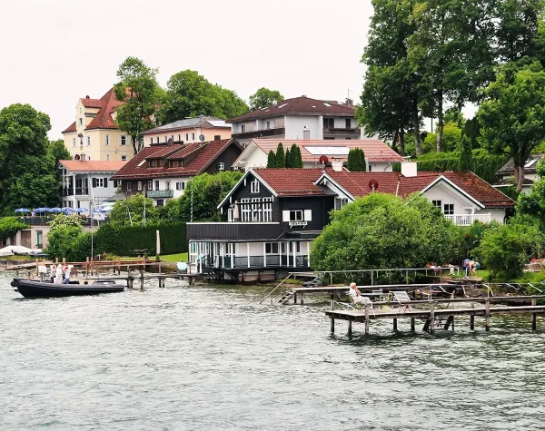 Häuser am Ufer des Starnberger Sees mit Booten und Stegen im Vordergrund, umgeben von üppigem Grün.
