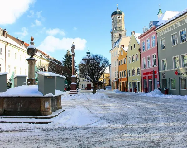 Verschneiter Stadtplatz in Weilheim-Schongau mit bunten Gebäuden und Kirchturm im Hintergrund.