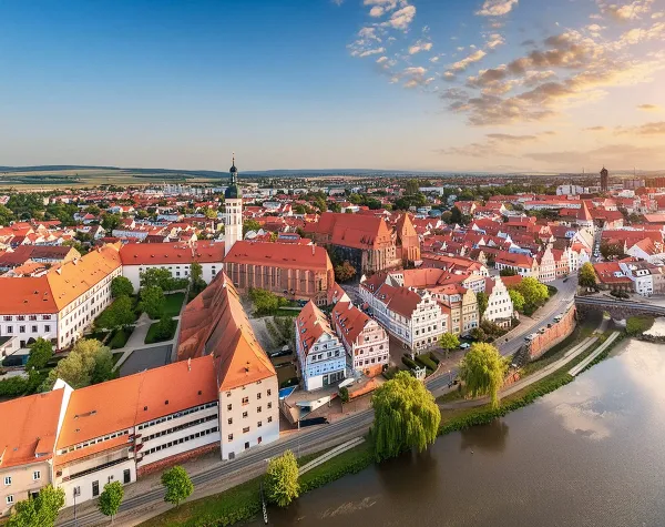 Panorama der Stadt Landshut mit roten Dächern, Kirche und Fluss im Vordergrund, umgeben von einer sanften Hügellandschaft.