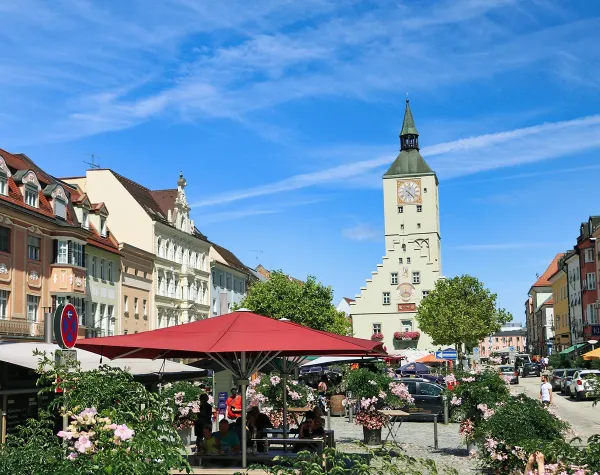 Historische Altstadt mit Straßencafés, blühenden Rosen und einem markanten Uhrenturm unter blauem Himmel in Deggendorf.