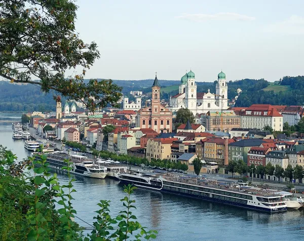 Blick auf Passau mit Kirche, historischen Gebäuden und Flusskreuzfahrtschiffen an der Donau bei sonnigem Wetter.