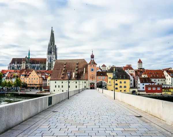 Steinerne Brücke in Regensburg mit Blick auf den Dom und bunte Altstadthäuser unter bewölktem Himmel.
