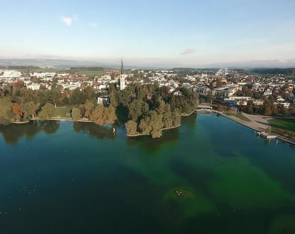 Luftbild von Cham mit Kirche im Zentrum, umgeben von Bäumen und Wasser, im Hintergrund städtische Bebauung.